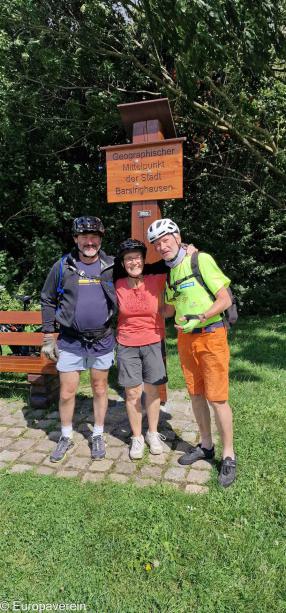 Zwei Männer und eine Frau in Fahrradkleidung stehen an einem Schild mit der Aufschrift "Geographischer Mittelpunkt der Stadt"