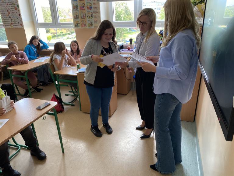 Das Foto zeigt einen Klassenraum in dem zwei Lehrerinnen vor der Tafel von einer Schülerin etwas gezeigt bekommen